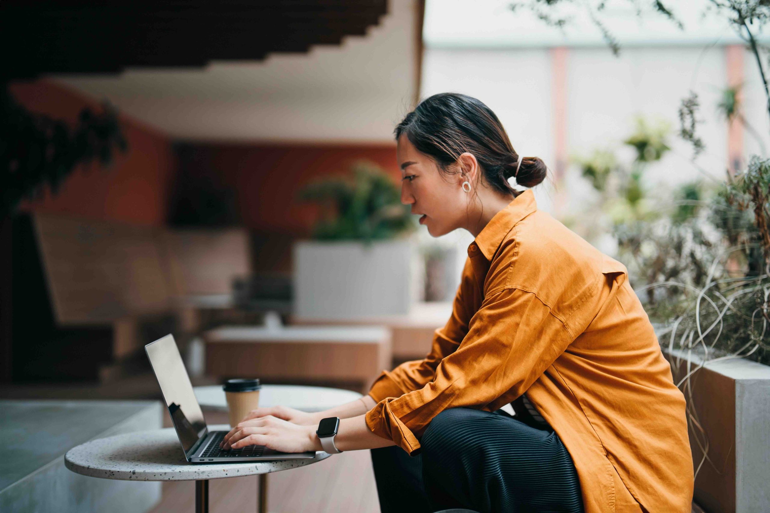 A woman in an orange shirt works on a laptop at an outdoor table.